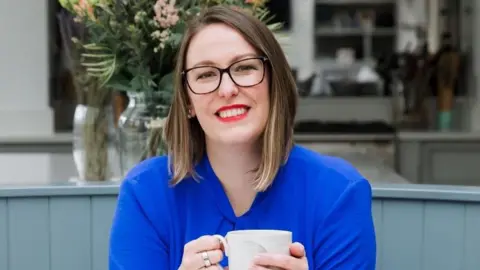 Yolanda De Vries A woman in a bright blue blouse is holding a cup of tea in a modern kitchen. She has dark glasses, brown hair and bright red lipstick.