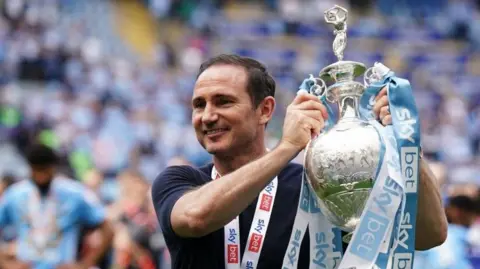 PA Media Coventry City head coach Frank Lampard holds up the Sky Bet Championship trophy following its presentation on the field after the final whistle following the Sky Bet Championship match at the Coventry Building Society Arena, Coventry. He has brown hair and is waring a navy short-sleeved top. He is smiling a he holds the trophy to his left. Blurred players can be seen on the pitch behind him, and supporters in the stands.
