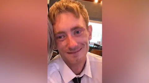 Conor Kinlan is a man with blond hair, wearing a white, collared shirt and a black tie. He is looking directly at the camera and is smiling.