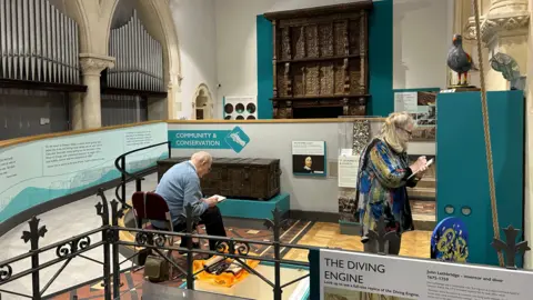 Two people draw in the museum. One is sitting sketching an antique chest, the other draws a small object in a display case.