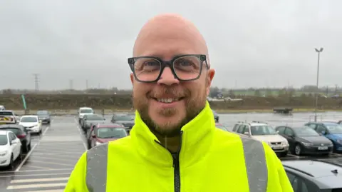 Tony Fisher/BBC A man in a yellow top looking into the camera with cars parked in a car park in the background. He is wearing glasses and has a short beard.