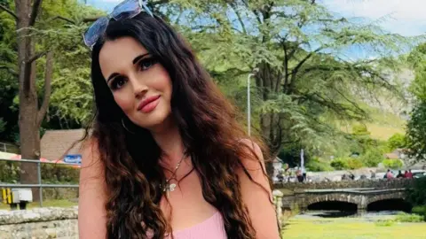 Diana Diana looking into the camera. She has sunglasses resting on her head and a small bridge and greenery can be seen in the background