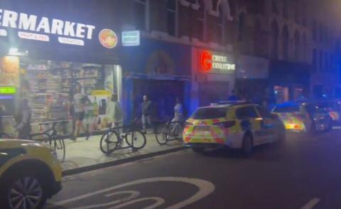 A photo taken at night shows a line of police cars parked up next to an independent supermarket as people walk past