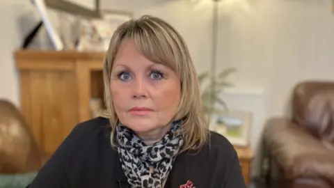 A women with blonde shoulder length hair is looking directly at the camera. She is wearing a black jumper and a leopard print scarf. She is sitting in a living room and you can see photo frames and a brown sofa in the background. 