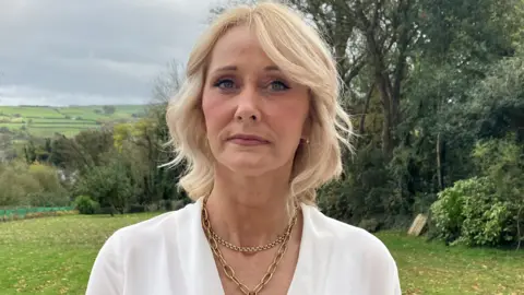 Deirdre Shakespeare wearing a white blouse and gold necklace. She has wavy blonde hair and is outside. There are trees and fields behind her.