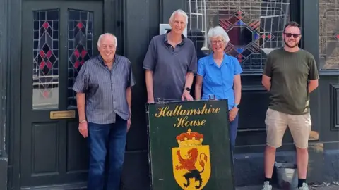 Four adults stand squinting in the sunshine outside a pub. The couple in the middle of the shot stand behind an old fashioned pub sign