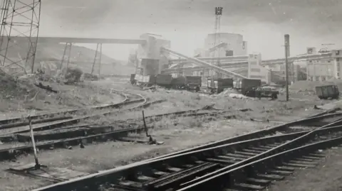 Chris Hockney A black and white image of rail tracks running through an industrial looking area with cranes, tricks and concrete-looking buildings.