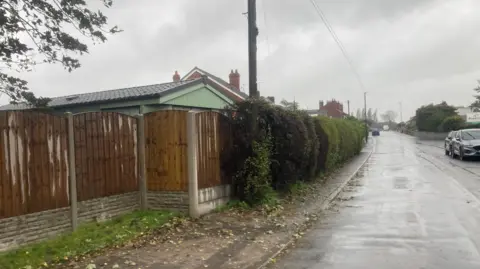 LDRS/Tony Gardner A residential road with a fence running down the left hand side. Behind the fence the top of caravans are visible with houses in the distance.