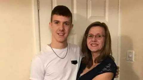 A mum and son are stood in a room looking at the camera in a white room. The man is wearing a white t-shirt and the woman a black dress. 