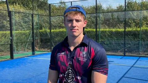 Charlie Stubbs / BBC There is a man with his back to camera wearing a blue backwards cap, a purple, pink and navy shirt. He has brown hair and is stood on a blue padel court.