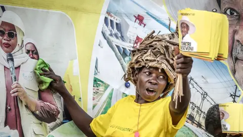 A supporter of Yoweri Museveni in a yellow campaign T-shirt holds flags with Museveni's face on them and is putting up posters with the other.