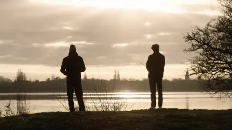 Max Morgan The silhouettes of two men fishing at a lake under a moody sky. The sun reflects off the water. There is a church spire in the distance.