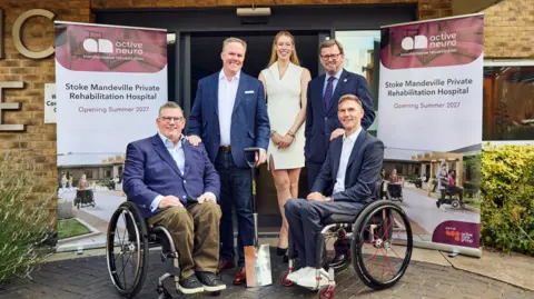Active Care Group Group of five people pose outside a building beside two banners reading “Stoke Mandeville Private Rehabilitation Hospital – Opening Summer 2027” and “active neuro transformative rehabilitation”, with two people seated in wheelchairs at the front and a ceremonial shovel placed between them.