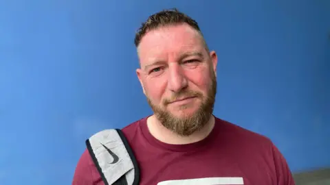 A man in a maroon t-shirt looks at the camera. Behind him is a blue wall. He has a grey backpack slung over his right shoulder. He has short hair and a beard.