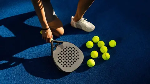 Close up of a player holding a padel racquets by a blue padel court on which are a number of yellow tennis balls
