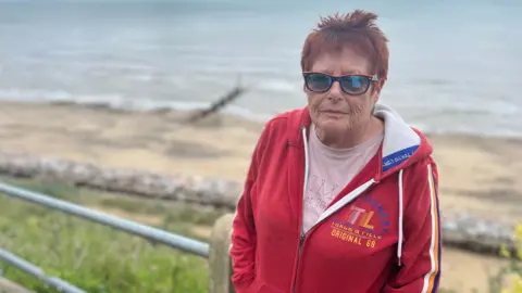 Jack Maclean/BBC A woman with sunglasses and a red jumper looks towards the camera from the top of a cliff above Mundesley beach, which can be seen in the background.