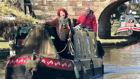 John Sadler and Sharon Wells navigating their way home on their narrowboat. Ms Wells has a brown jumper and green scarf and Mr Sadler is wearing a red coat. They have a dog on the boat. There are other narrowboats behind them on the canal. The pair are looking happy in the sunshine.