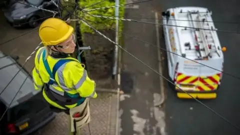 KCOM A woman in a hi-vis vest and wearing a helmet and a utility belt up a telegraph pole. There is a van with yellow and red chevrons painted on its back doors parked on the road below.