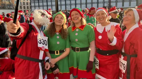 Five people, three dressed as Santa and two as elves, smile into a camera above them on a selfie stick. Behind them more Santa's can be seen dressed up in the square. 