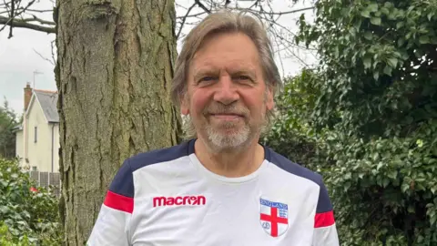 Shivani Chaudhari/BBC A man wearing a football shirt and smiling at the camera. He is standing outside next to a tree.