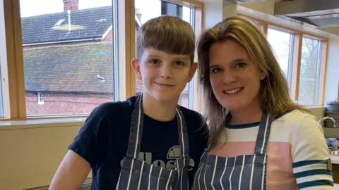 Jools, a young teenage boy with short dirty blond hair and a fringe, and his mum, Ellen, a woman with a similar hair colour whose hair sits on her shoulders, smile as they stand in what appears to be a school kitchen with navy and white striped aprons on.