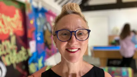 A woman with short blonde hair and blue glasses is smiling at the camera. She is wearing a black top.