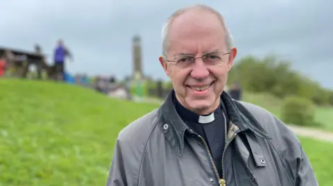 Justin Welby, wearing a wax coat on top of his usual clerical outfit, smiles as he stands near the Crich Memorial tower in Matlock, Derbyshire