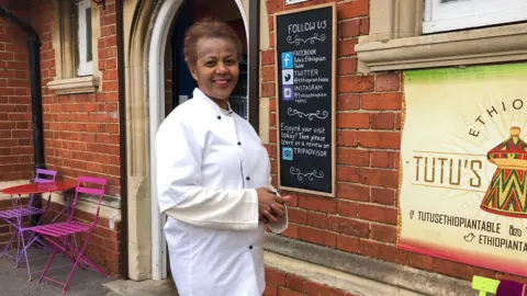 Tutu Melaku in a chef's tunic standing outside the door to her Ethopian cafe. She is smiling. The cafe is housed in a red brick Victorian building. There is a pink outdoor cafe table and chairs to one side of the door, to the other side there is the restaurant sign showing a traditional Ethopian cooking pot, and a blackboard advertising the cafe's social media sites.
