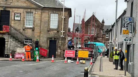 Charles Brown A picture of road works in Sanquhar with scaffolding out on the roadway next to the historic Tolbooth Museum on the left. There is a road closed sign and traffic lights and a man in orange overalls on the phone and another in a high-visibility jacket and hard hat talking to a member of the public
