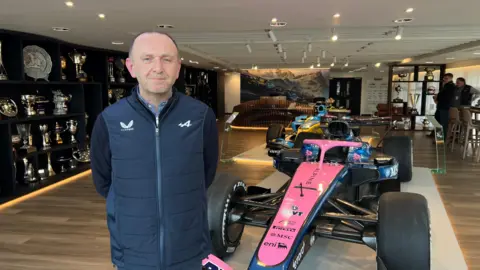Ian Pearce, operations director at Enstone-based Alpine, posing for a picture in front of one of their cars. Trophies can be seen in a glass cabinets behind him. Two people can be seen looking at the cars in the far right corner of the room.
