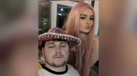 Charlie Hurst Man in a pink cowboy hat, Charlie, takes selfie with girl in a pink wig, Áine. They appear to be at a fancy dress party.