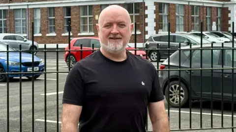 Barney Harte A bald man with a short white beard smiles close to the camera. He is wearing a black t-shirt with a small white logo on his left chest. Behind him is a black gate, with cars parked outside a red brick building with white corners.