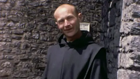 Father Thaddeus Kotik stands successful  beforehand   of the walls of Pembroke Castle. He is wearing a achromatic  habit. He is bald, with a small  acheronian  hairsbreadth  connected  the sides of his head. He is smiling into camera. 