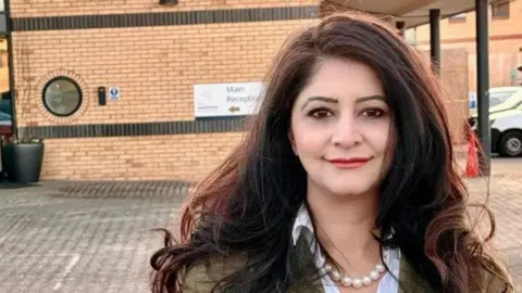 Peterborough Labour Dr Shabina Qayyum stands in the car park of a doctor's practice. She has dark flowing hair and is wearing a green jacket, pearls and a shirt.