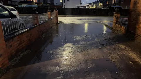 LDRS A large pool of water reflects streetlights and spans a domestic dirt-track lane between two brick walls 