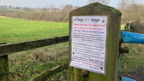 Elizabeth Baines / BBC A sign discussing dog waste bags is pinned to a post in the country side. Rolling hills are behind.
