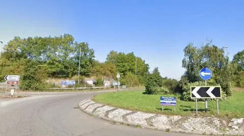 Google A grassed roundabout with trees and shrubs in its middle on the A45 with a white sign to the left saying Mawsley, Old and Lamport and below a brown heritage saying Lamport Hall