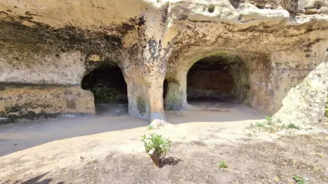 Janine Tanner Two caves, cut into pale sandstone cliffs, with a level area in the foreground
