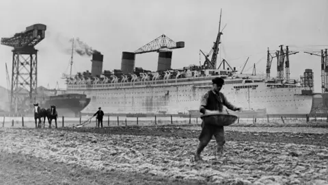 Getty Images A black and white image of a large ocean liner with a large crane in the left of the shot and people sowing fields in the foreground