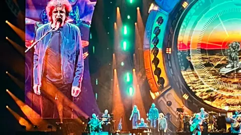 Jeff Lynne on stage singing into a microphone, stood with band members with a vibrant multi coloured backdrop. A screen is showing a larger version of Lynne to the right of the stage