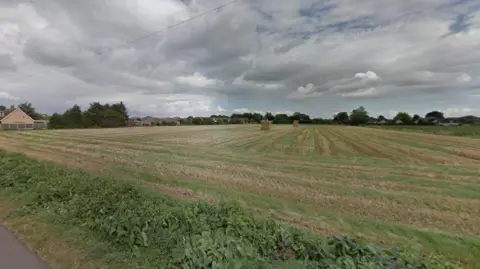 A field with hay stacks. A number of bungalows are on the left hand side and trees are around the edge of the field. 