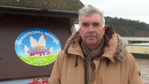 Robert Phipps looking at the camera and stood in front of an ice cream hut on a seafront promenade. 