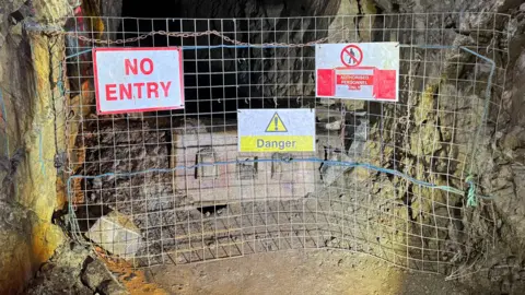 A square metal fence blocks off a mine shift. Three signs are on the fence - one says no entry, with red letters on a white background. Another, in the middle, says danger in black writing on a yellow background, with the danger symbol of an exclamation mark in a triangle. The final signs says no unauthorised personnel. 