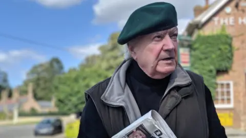 Tony Martin wearing a beret and coat, clutching a folder and a newspaper. He is standing outside a property covered in ivy