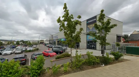 Google Google Street View image of Pure Gym - a large white building - and the car park. There are some small trees in the foreground and the car park appears to be full.