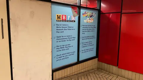The closed ticket office. The door has been sealed up with plyboard. Posters, advertising the services previously offered at the ticket office, remain on the glass windows but are pealing due to their age.