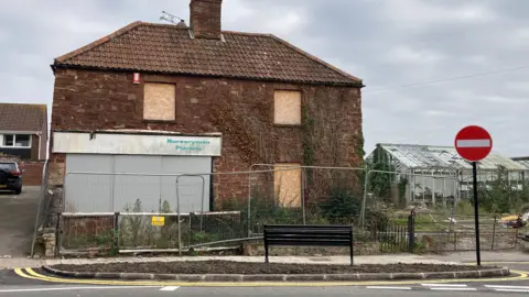 The view from across the street, capturing a black bench overlooking a derelict square brick cottage with boarded up windows and metal fencing surrounding the front garden. On the right of the house there is an abandoned greenhouse with smashed glass and overgrown greenery.