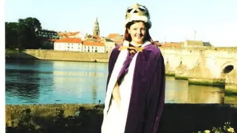 Amy Campbell A young woman stands by the river with the Old Bridge pictured behind her. She has dark hair and is wearing a purple gown and a crown 