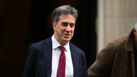 Ed Miliband wearing a navy suit with a white shirt and a red tie. He is smiling.