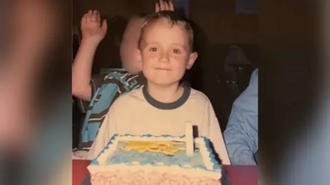 Ben Cole Edwards Photograph of Ben Cole Edwards sat in front of his pokemon birthday cake at the forefront. He has brown eyes and brown hair and smiles at the camera. A young boy can be seen behind him, however his face is blurred. Ben wears a white t-shirt with a navy trim collar. 